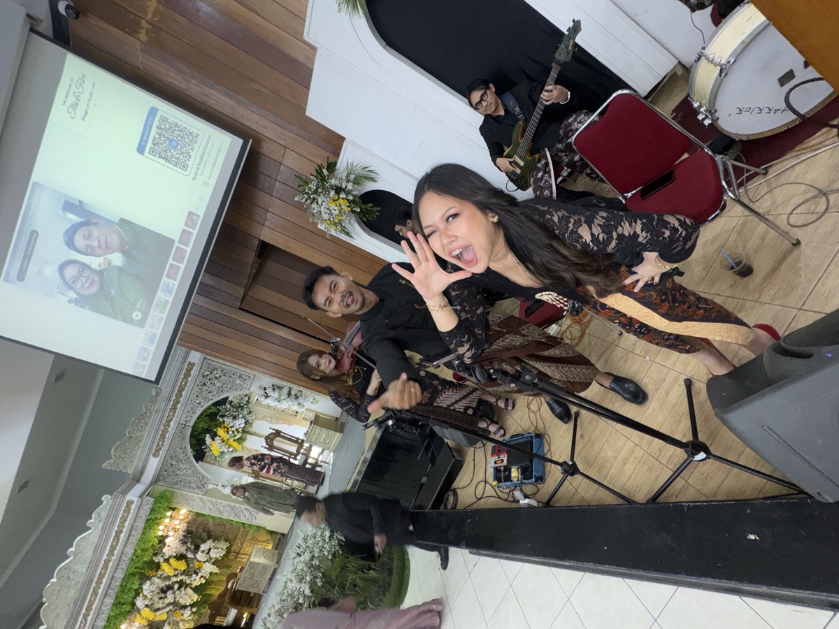 Penyanyi band menunjuk layar proyektor yang menayangkan foto tamu dan QR Berbagi Foto